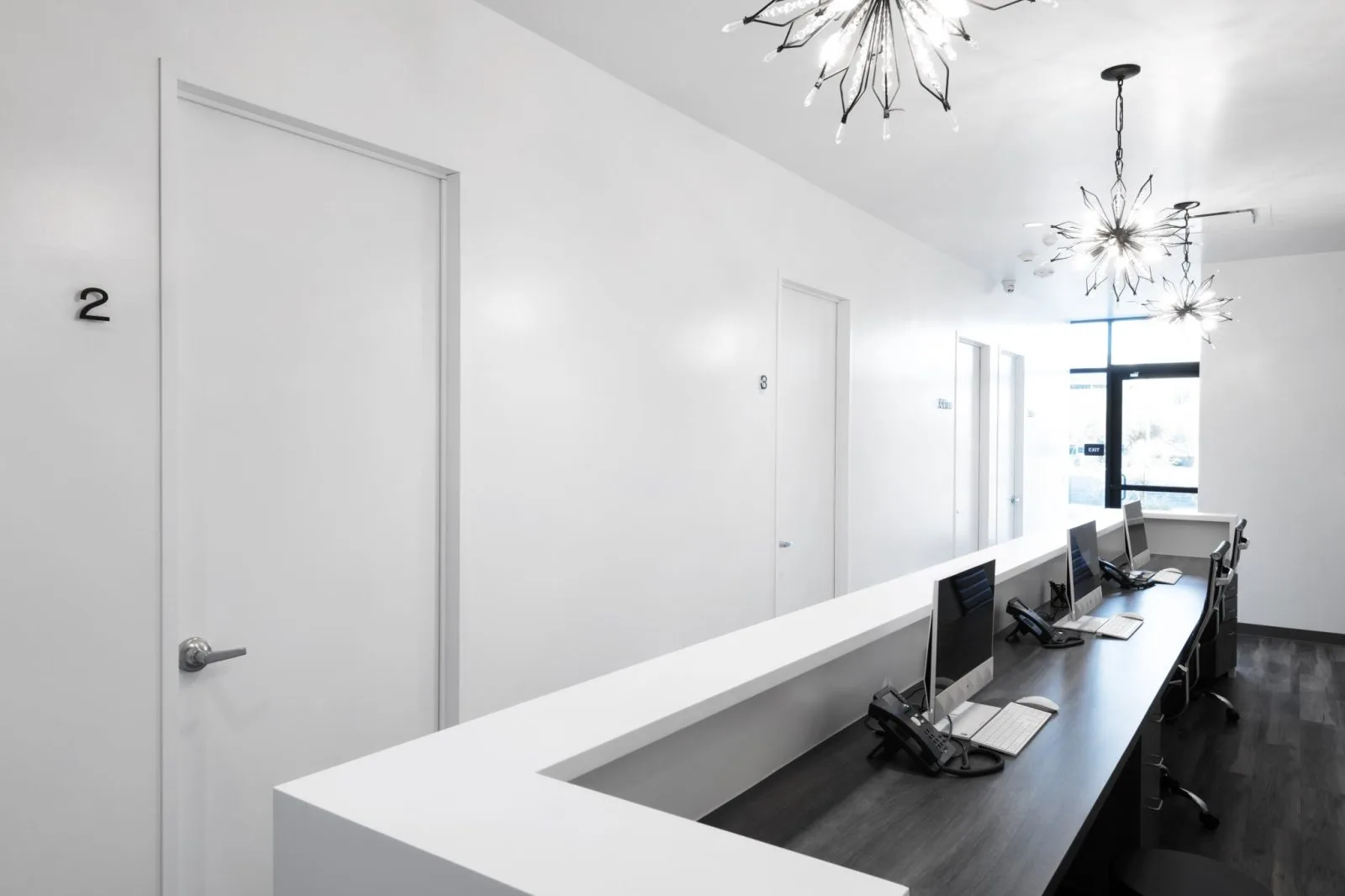 Modern reception desk with white counter, mirrored wall, and multiple exam room doors in a bright clinical hallway